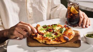 A person holding a slice of pizza on a wooden board, with a glass of iced beverage and a small bowl of sauce on the table.