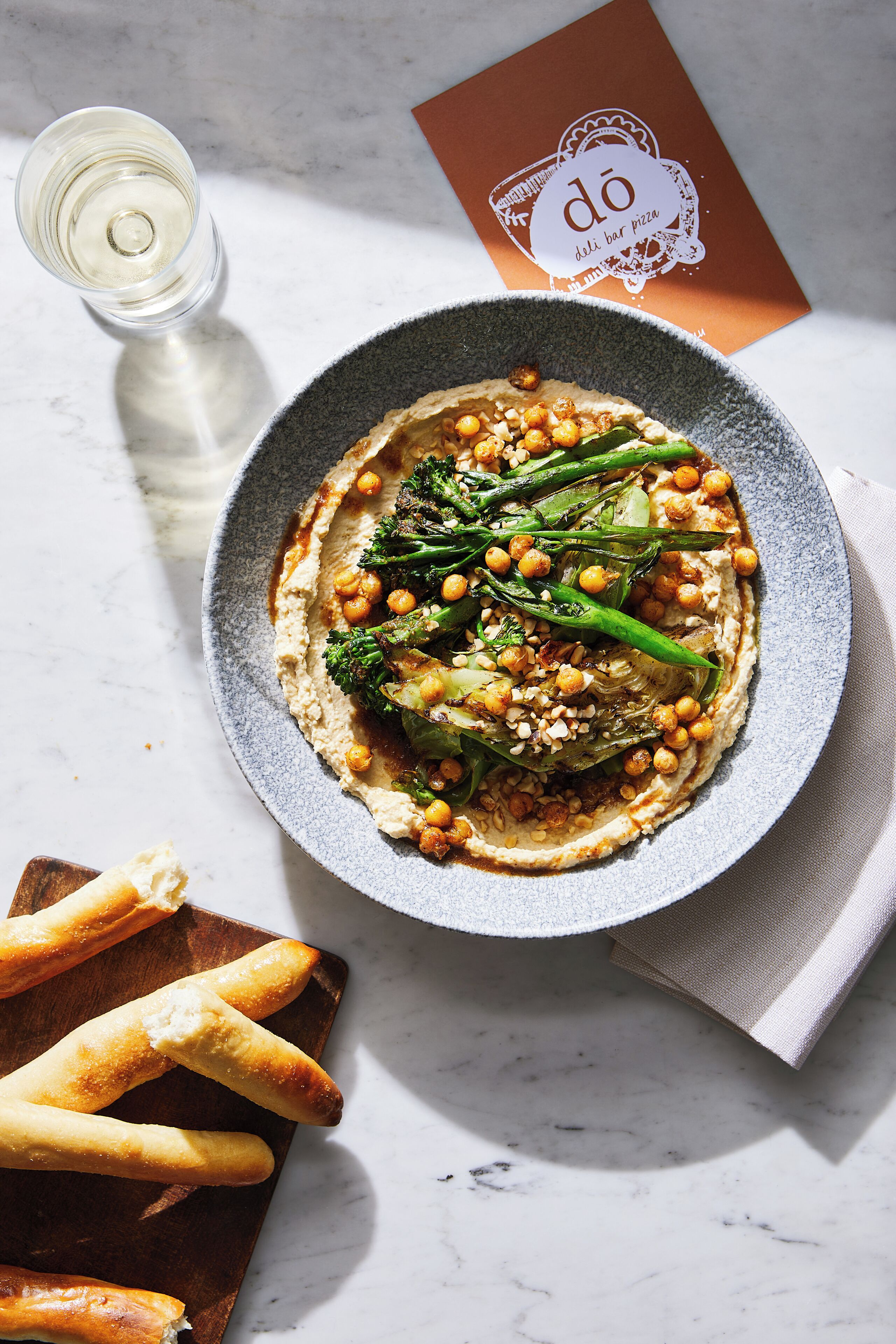 Une assiette de nourriture comprenant un bol de houmous, des légumes rôtis et des pois chiches, accompagnée d’une bouteille d’eau et d’une serviette en papier, le tout posé sur une surface en marbre.