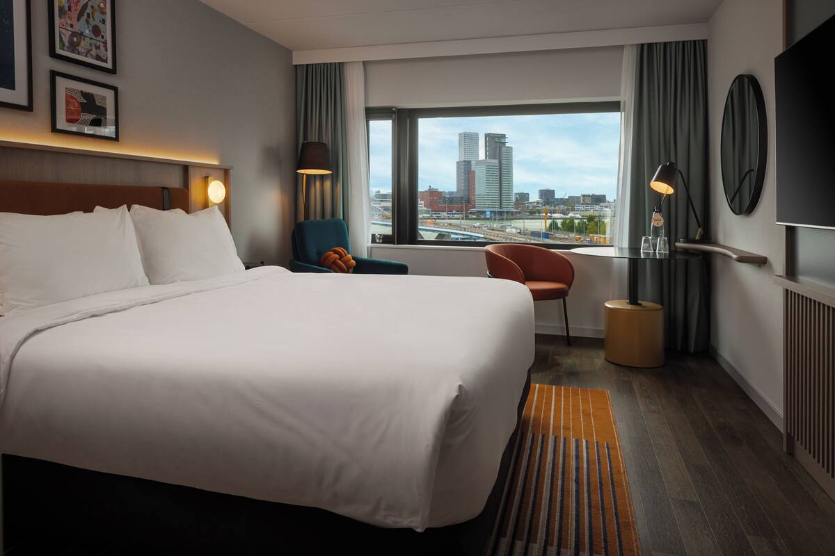 A hotel room with a large bed, a view of the cityscape, a desk with a lamp, and a chair.