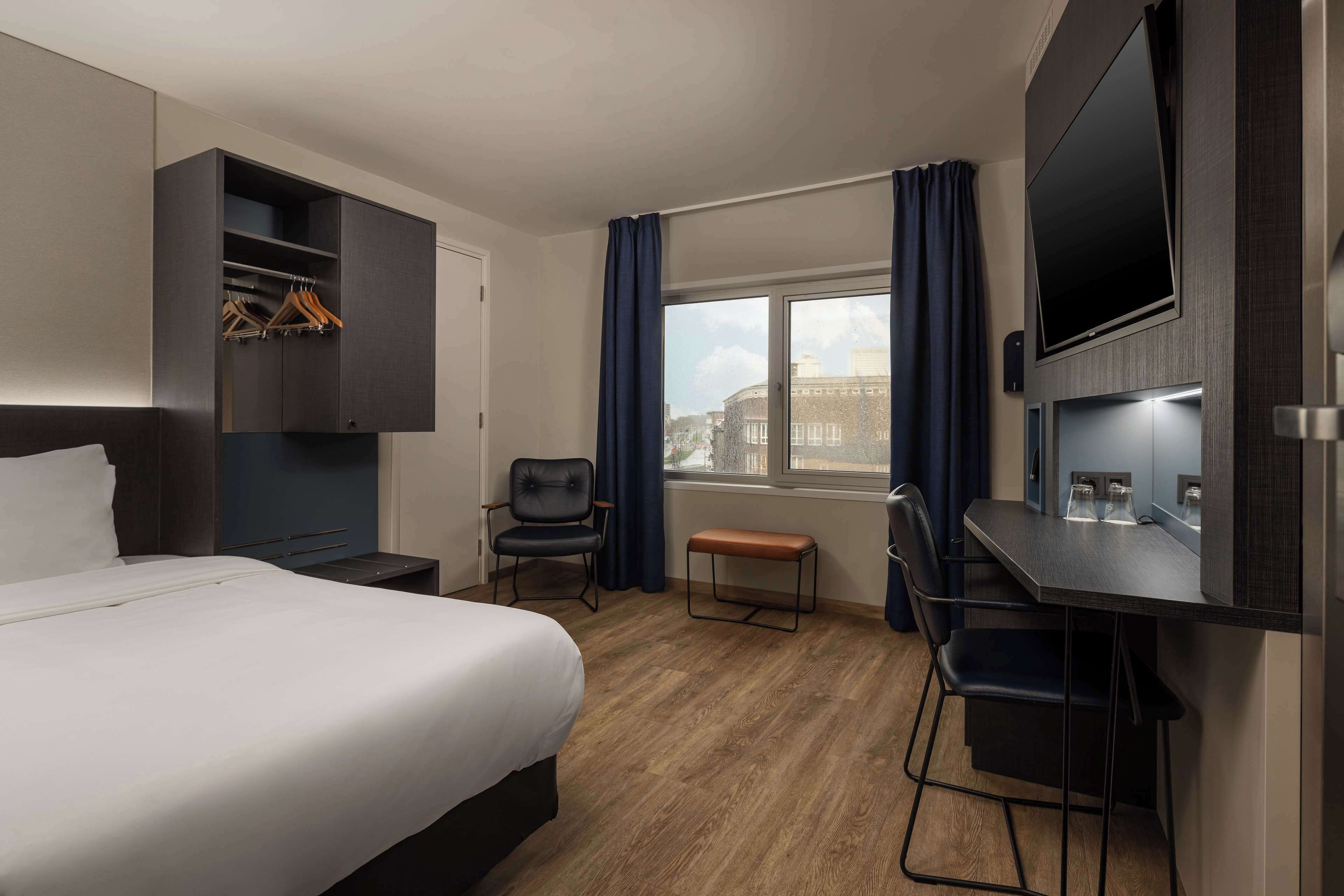 Une chambre d’hôtel comprenant un lit, un bureau avec une chaise, un téléviseur fixé au mur et une fenêtre avec des rideaux.