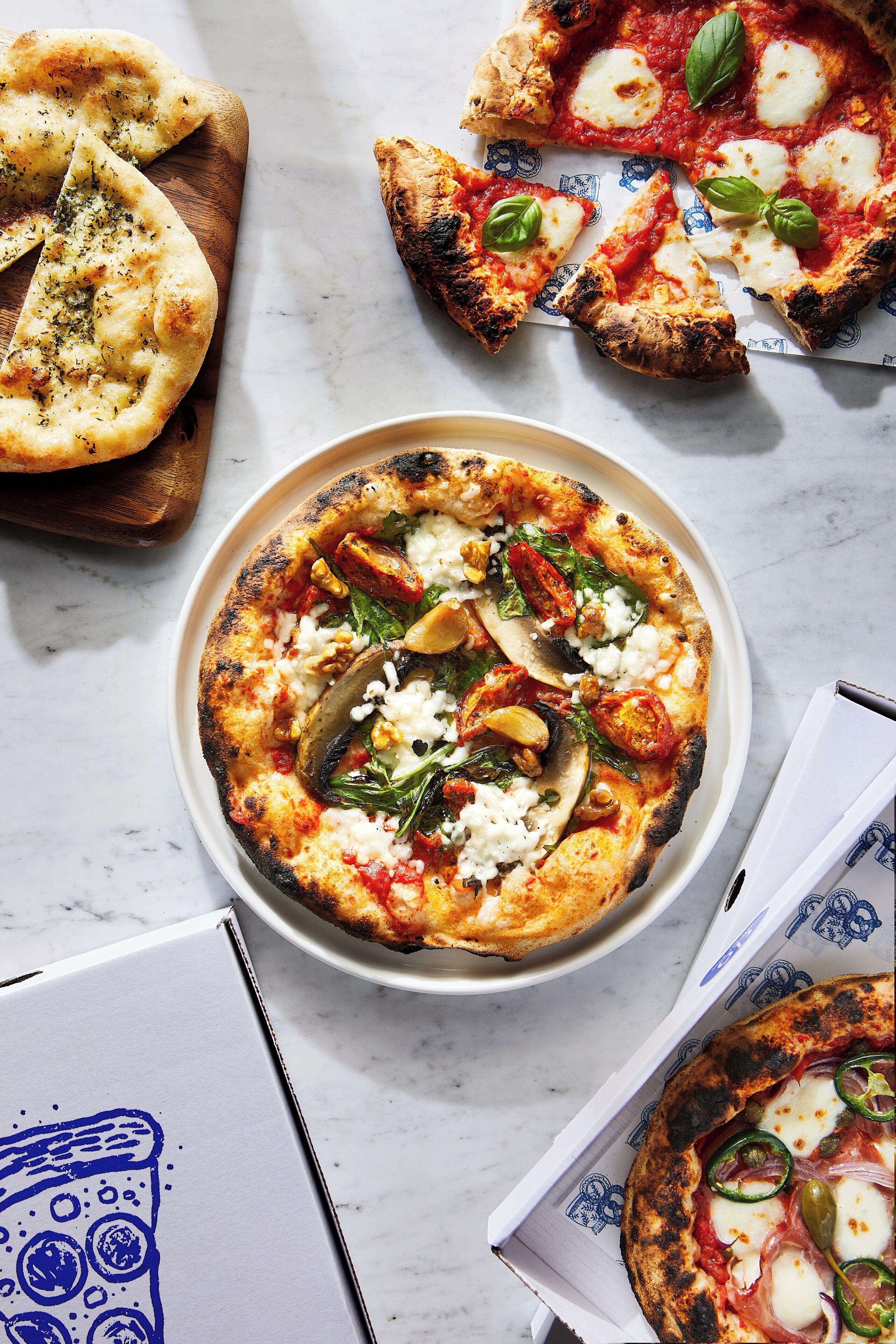 Pizza com diversos ingredientes, uma tábua de madeira com flatbread e caixas de pizza.