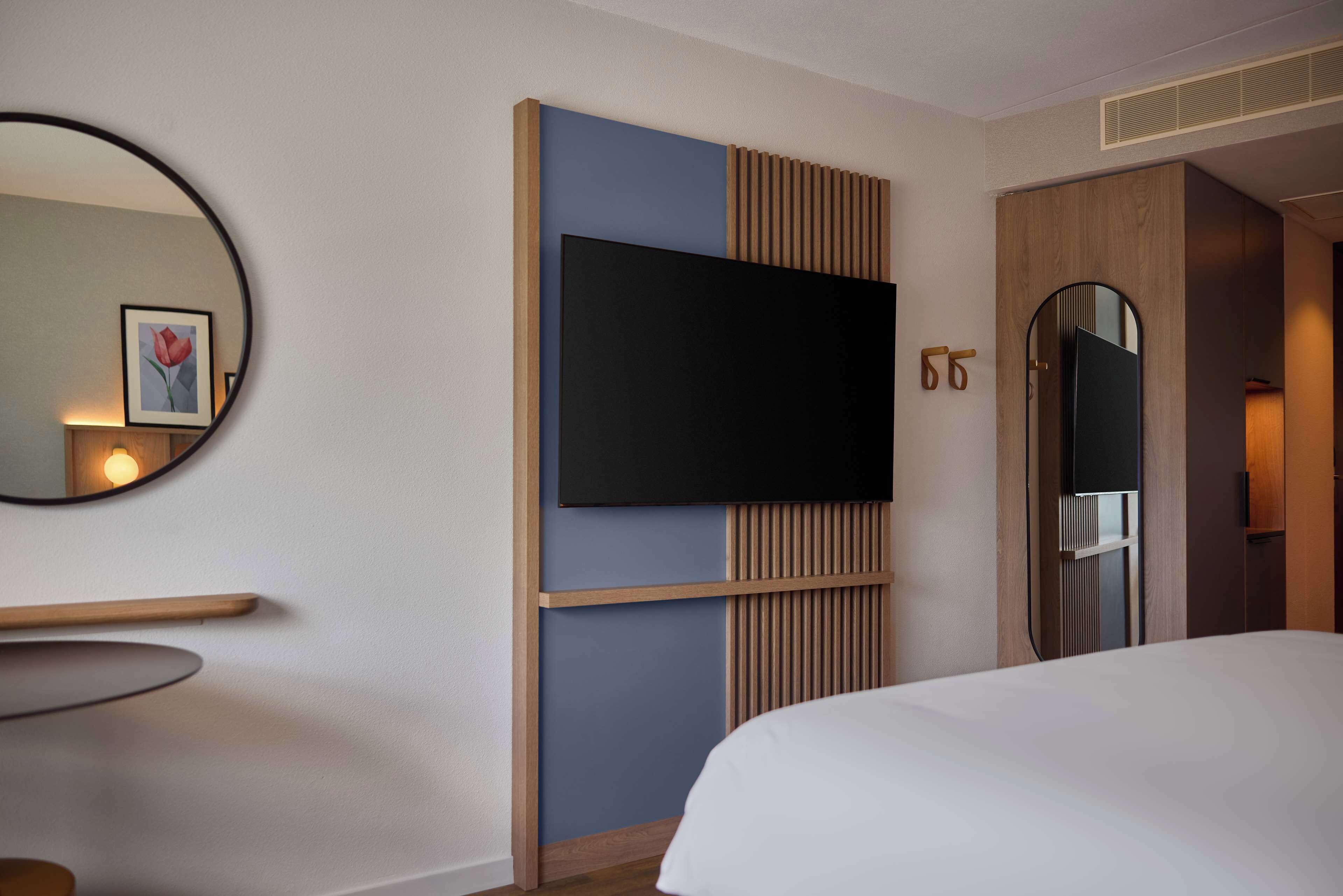 Une chambre d’hôtel avec un grand lit, un téléviseur fixé au mur, une armoire en bois, un miroir et un tableau encadré accroché au mur.