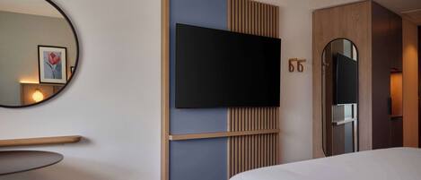 Une chambre d’hôtel avec un grand lit, un téléviseur fixé au mur, une armoire en bois, un miroir et un tableau encadré accroché au mur.