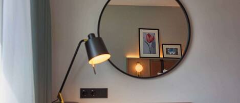 A compact room with a round table, a single yellow chair, a desk lamp, and a wall-mounted mirror reflecting a framed picture and a small lamp.
