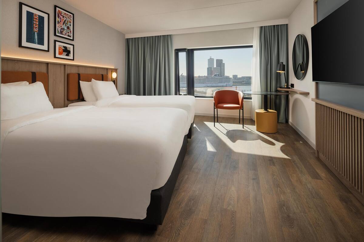 A hotel room with two beds, a large window with a city view, a chair, and a TV.