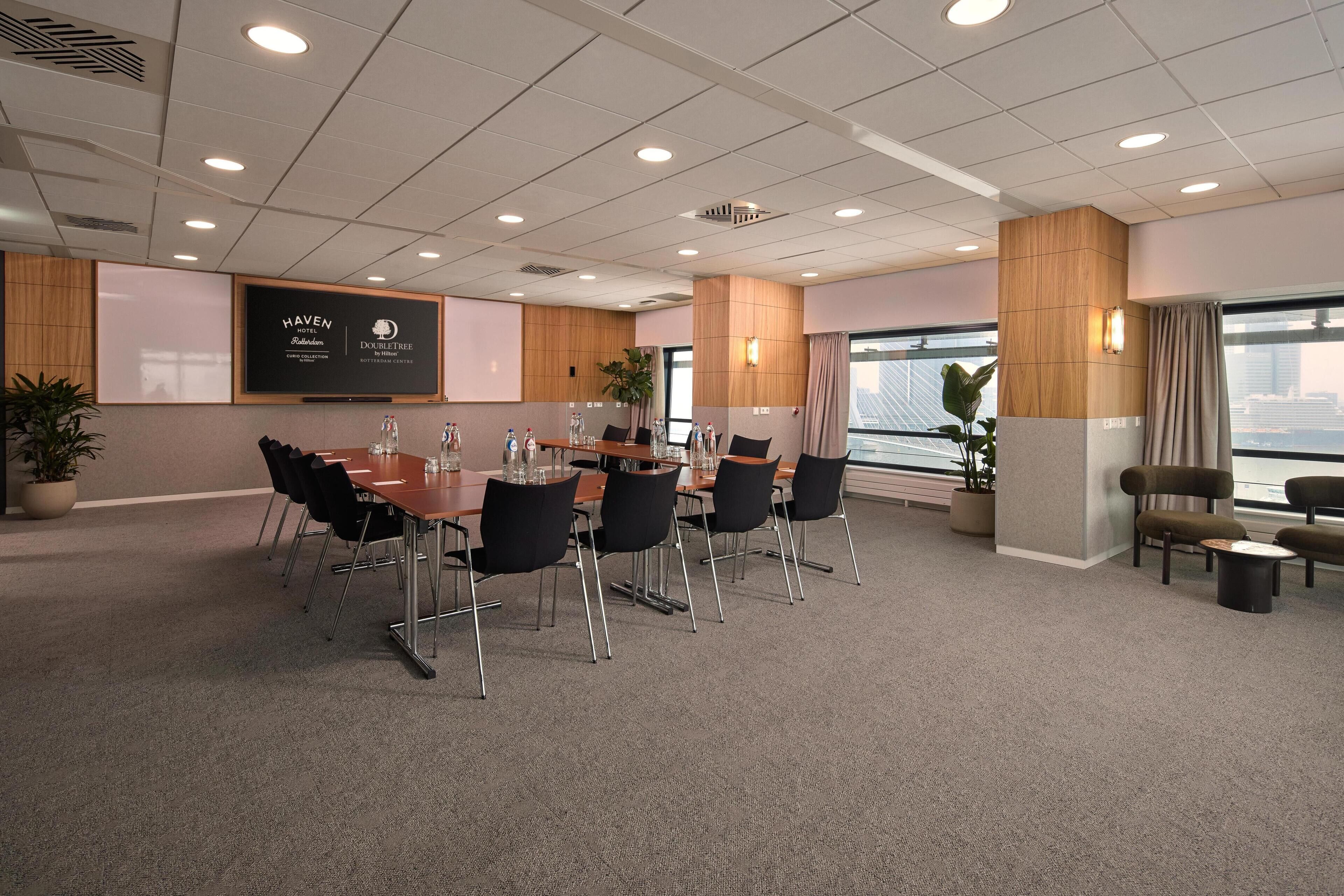 A conference room with a long table, black chairs, and a large screen displaying the hotel name.