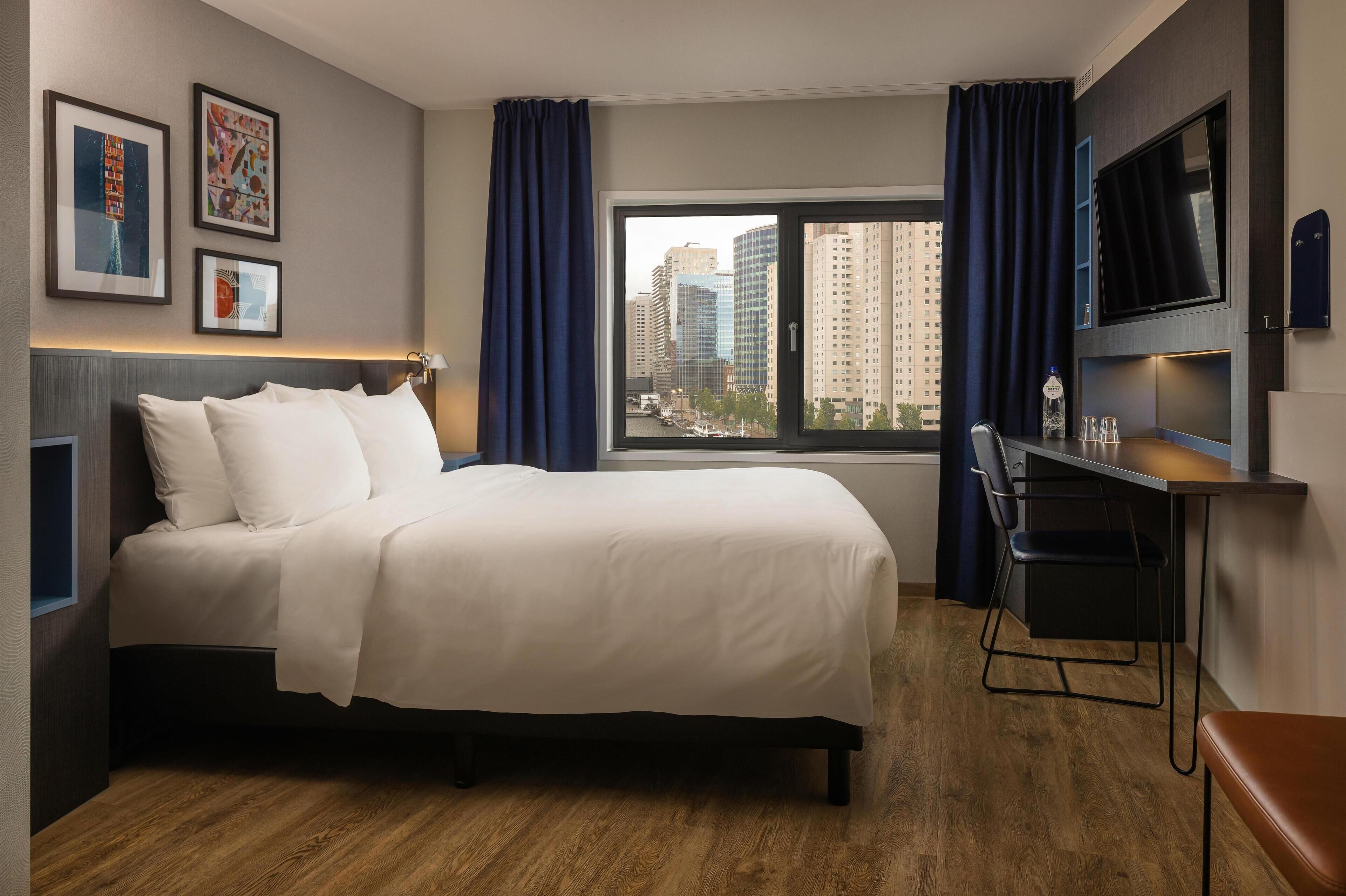 A modern hotel room with a large bed, a desk with a chair, a TV mounted on the wall, and a window with curtains.
