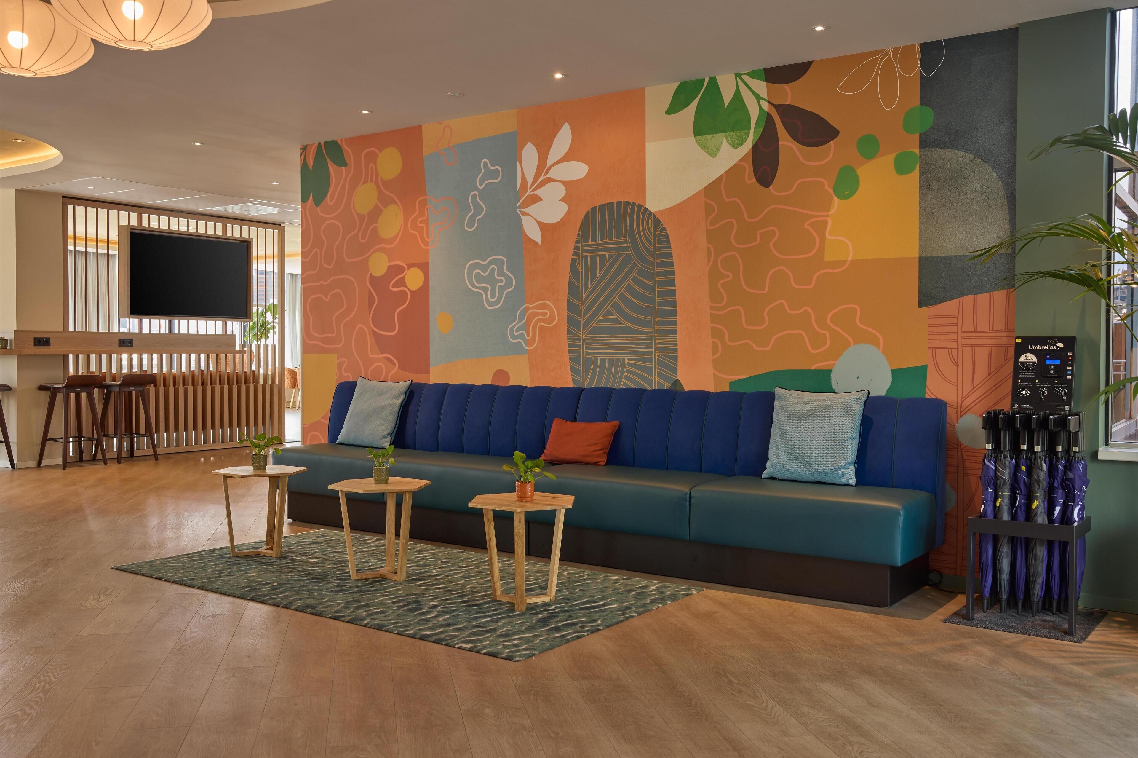 Un espace de détente moderne avec un canapé bleu, des tables en bois et une fresque murale abstraite aux couleurs vives.