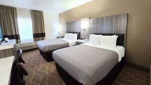 A hotel room with two beds, a desk, and a chair. There is a window with curtains, a wall-mounted lamp, and a ceiling light.