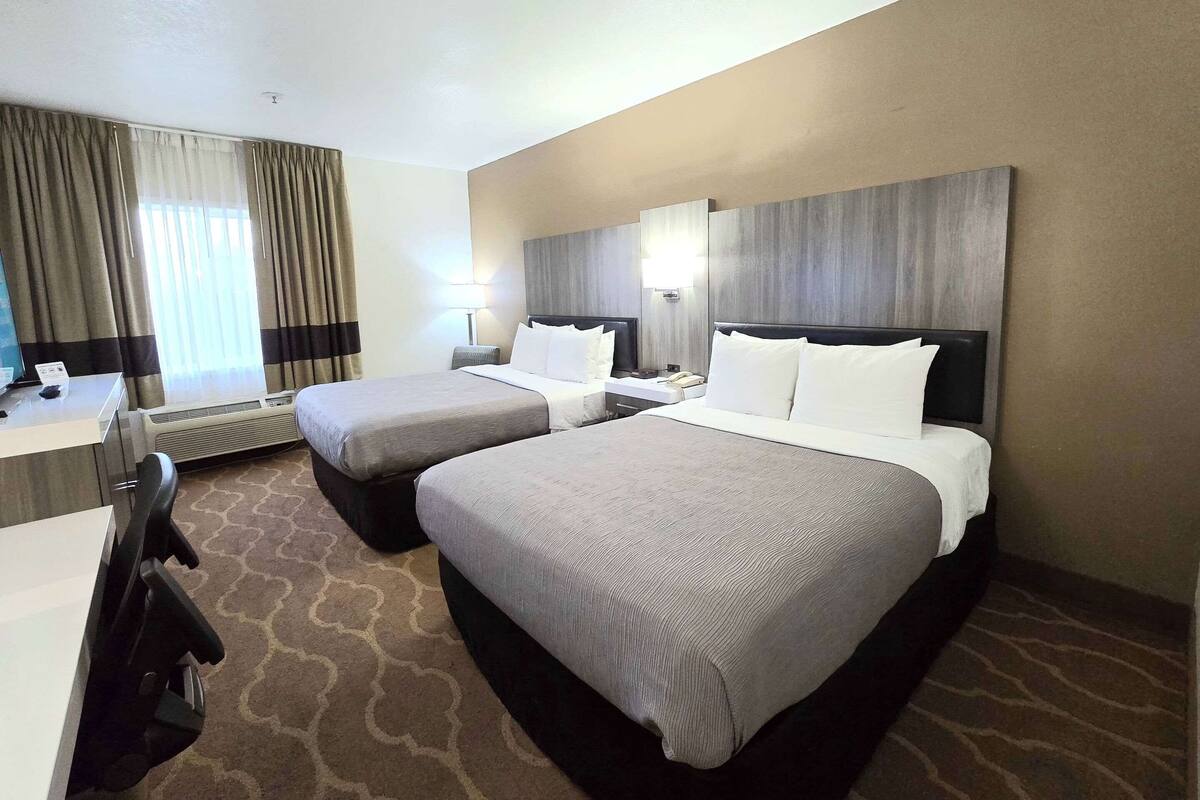 A hotel room with two beds, a desk, and a chair. There is a window with curtains, a wall-mounted lamp, and a ceiling light.