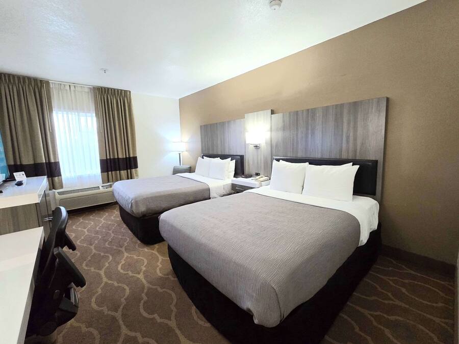 A hotel room with two beds, a desk, and a chair. There is a window with curtains, a wall-mounted lamp, and a ceiling light.