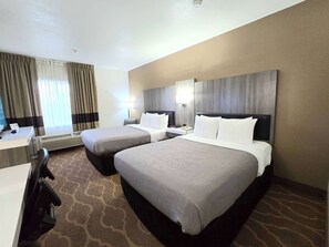 A hotel room with two beds, a desk, and a chair. There is a window with curtains, a wall-mounted lamp, and a ceiling light.
