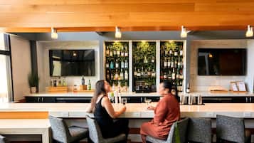 Uma área de bar com um balcão comprido, banquetas, uma TV na parede e prateleiras com garrafas.