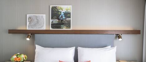Una cama bien hecha con sábanas blancas, una almohada anaranjada como detalle y dos cuadros enmarcados encima.