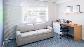 Ein Zimmer mit einer Couch, einem Schreibtisch mit Stuhl, einer Stehlampe und einem Fenster mit Jalousien.