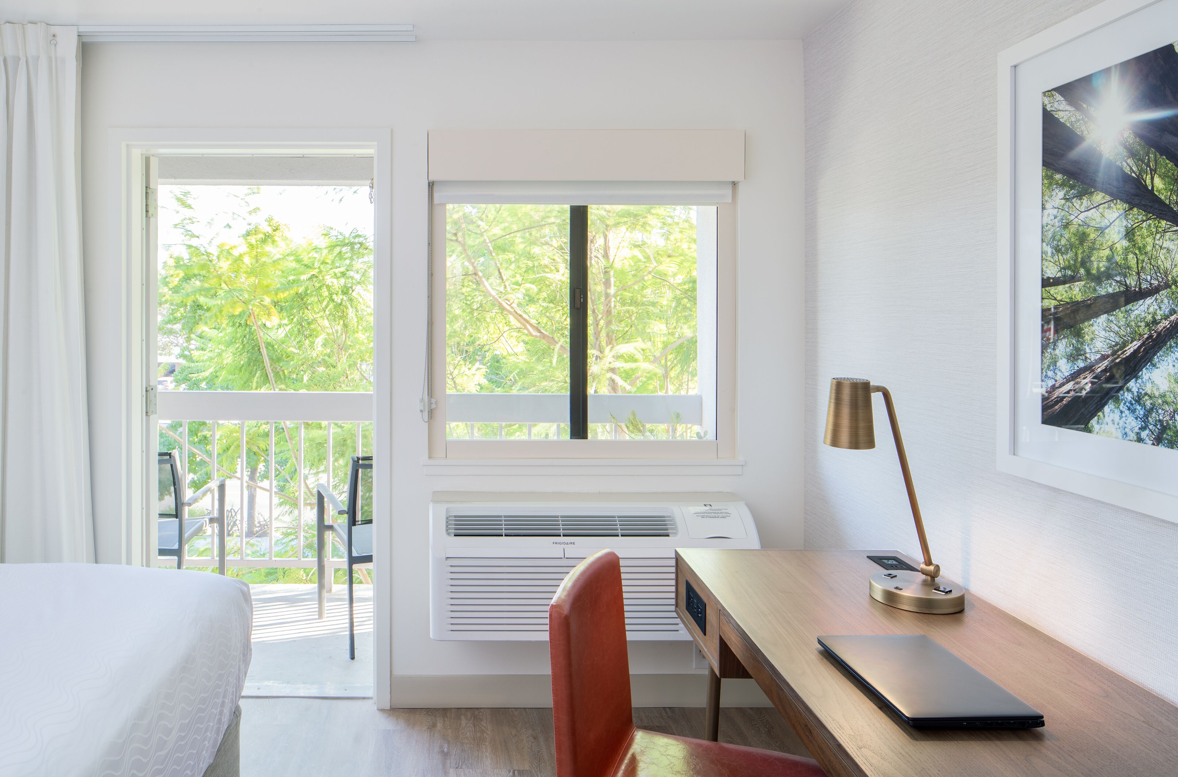 A hotel room with a bed, a desk with a laptop, a chair, and a window with a view of trees.