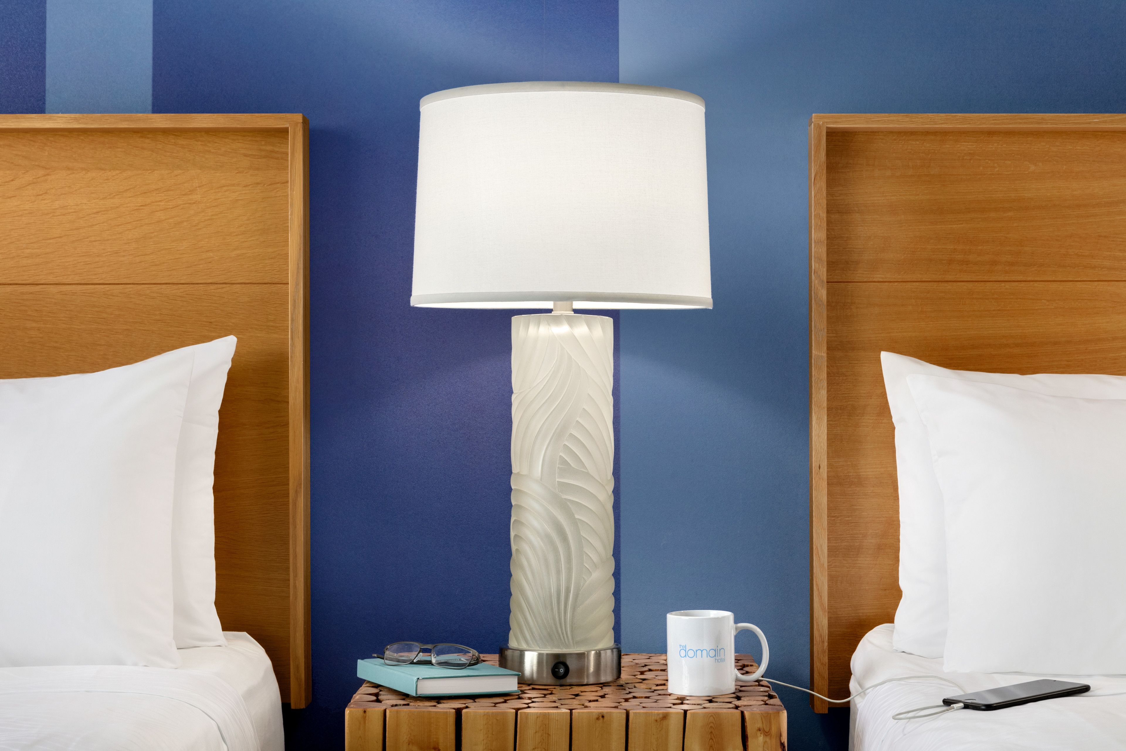 A hotel room with two beds, a nightstand with a lamp, a mug, a book, and a smartphone.