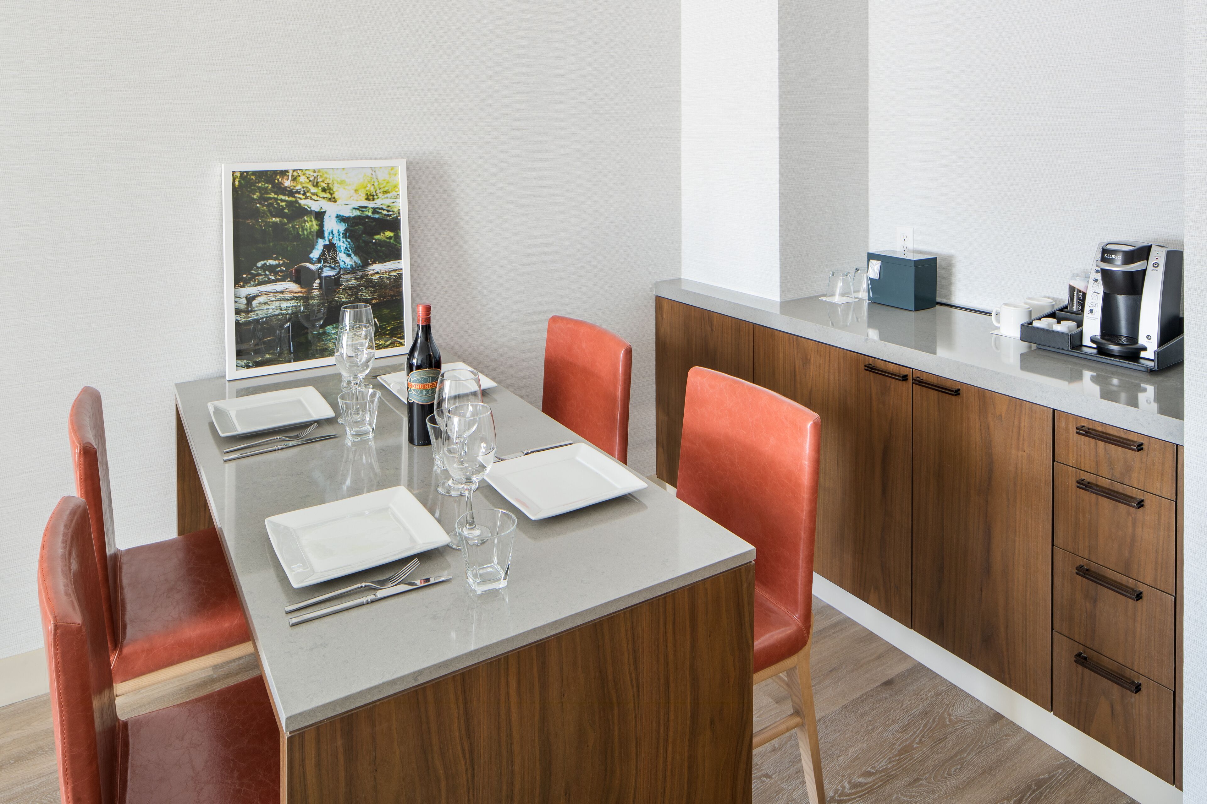 Comedor con una mesa dispuesta para cuatro personas, equipada con copas de vino, platos y cubiertos. Hay una cafetera y una pequeña caja azul en la encimera.
