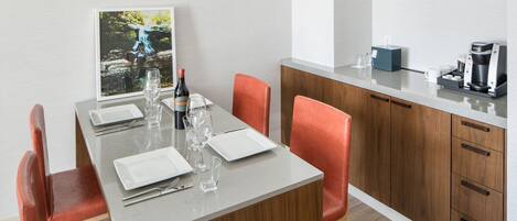 Comedor con una mesa dispuesta para cuatro personas, equipada con copas de vino, platos y cubiertos. Hay una cafetera y una pequeña caja azul en la encimera.