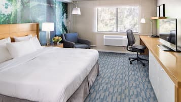 Quarto de hotel com uma cama grande, uma escrivaninha com computador, uma cadeira, um abajur e uma janela ampla com persianas.