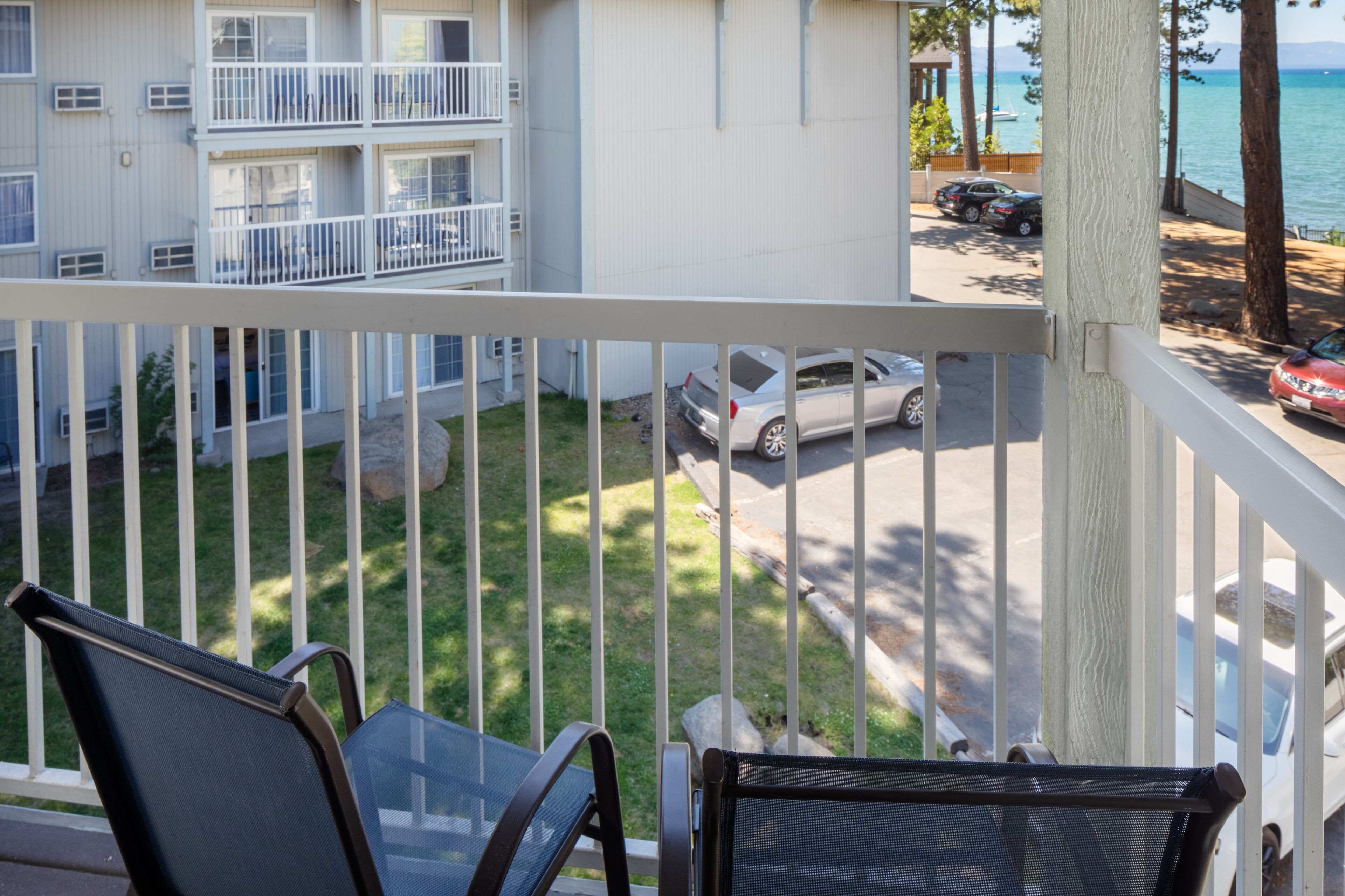 A balcony with a view of a multi-story building, a parking area with cars, and a body of water in the background.