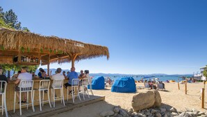 Un bar situé en bord de mer, avec des tables à l’extérieur et des parasols à toit de chaume.