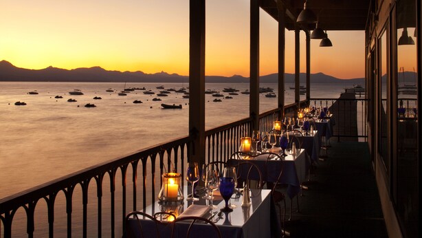 Un restaurant avec des tables dressées pour le service, donnant sur une étendue d’eau où se trouvent des bateaux.