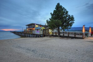 Un edificio frente a la playa con una terraza de madera que se extiende sobre el agua, un letrero que indica un restaurante y un gran árbol cercano.