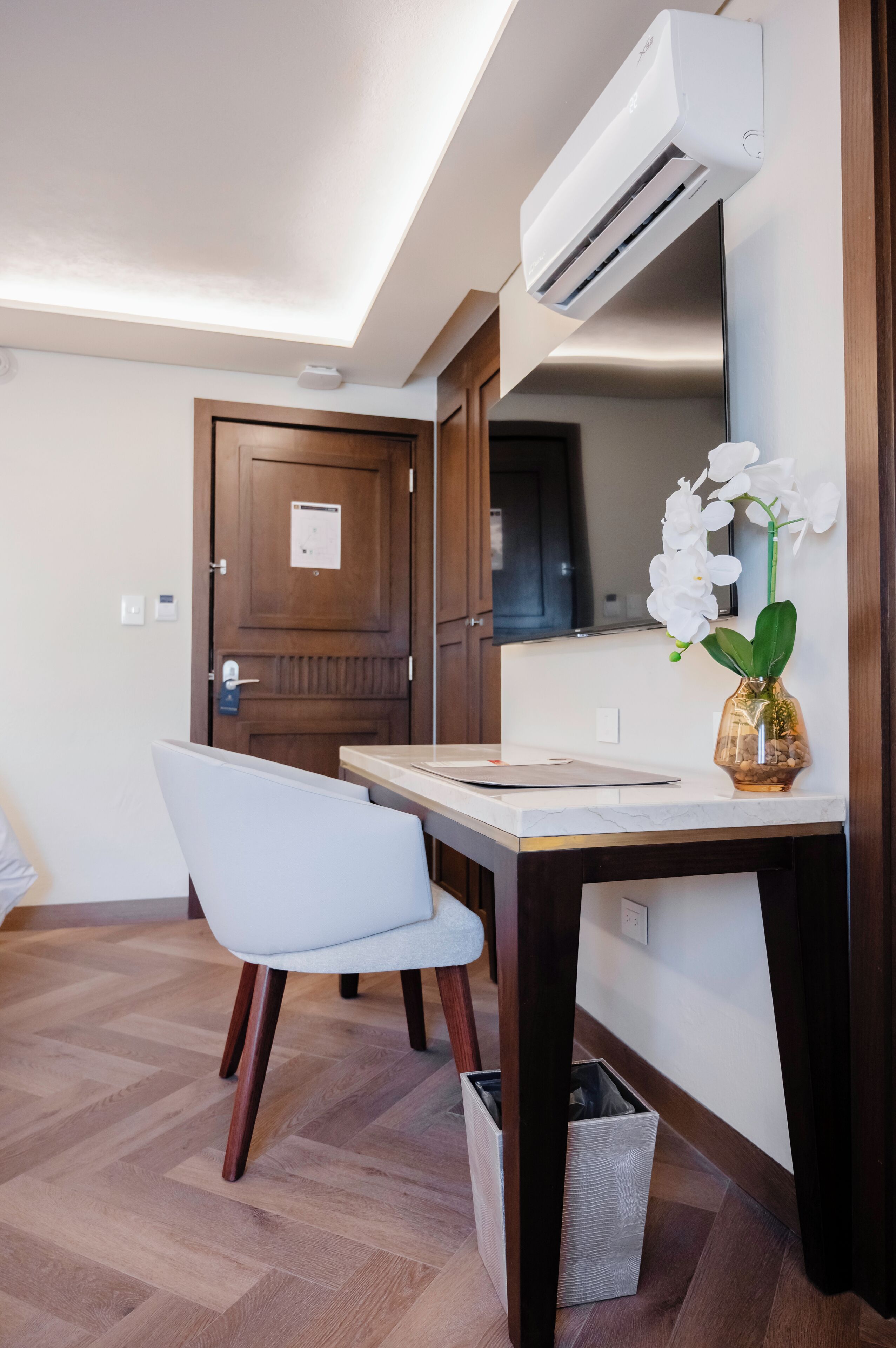 Una habitación moderna con suelo de madera, una silla blanca, un escritorio, un aire acondicionado blanco, una puerta de madera y un jarrón con orquídeas blancas.