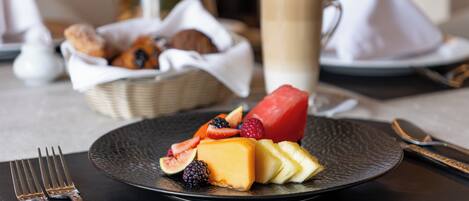 Een ontbijtsetting met een bord kaas, fruit en een mand met croissants op de achtergrond.