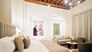 A modern hotel room with a large bed, a sofa, a chair, a small table with a vase, and a chandelier. - Quinta Real Puebla (Puebla)
