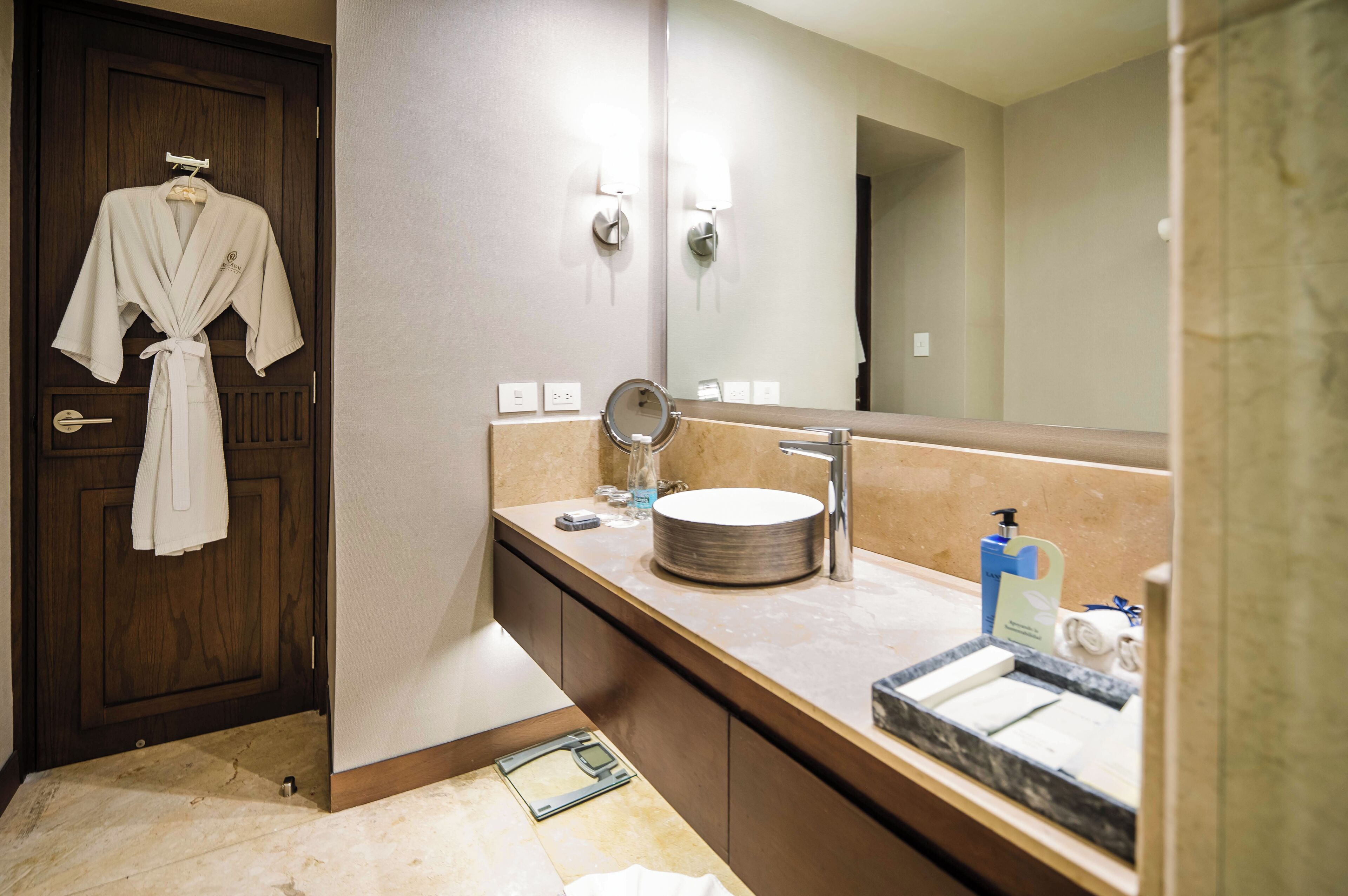 Un baño con puerta de madera, una bata colgada en la puerta, un espejo redondo, un lavabo y artículos de aseo en la encimera.