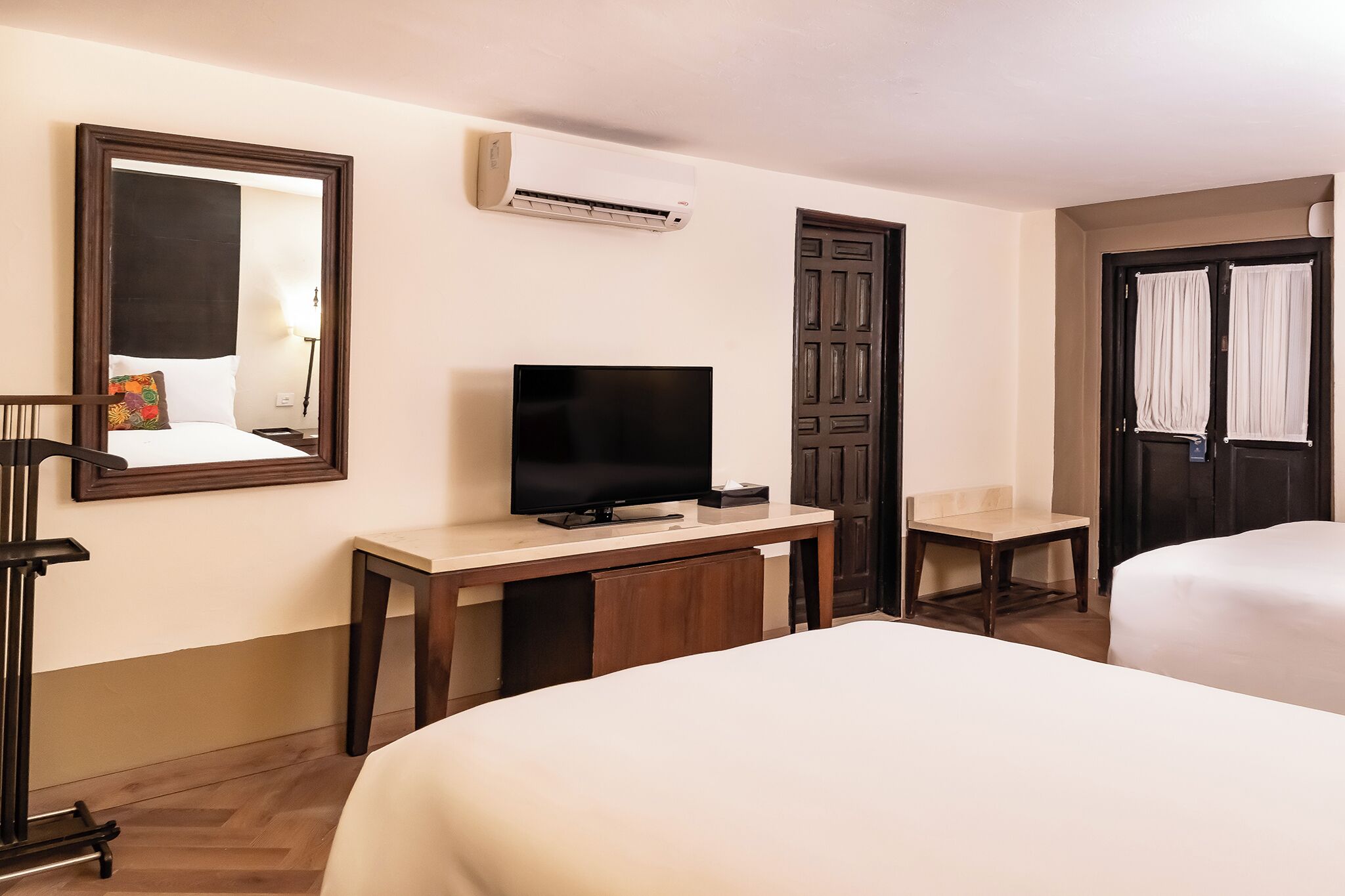 A hotel room with two beds, a TV, a desk, a chair, an air conditioning unit, and a mirror.