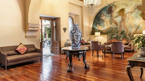 A spacious room with a wooden ceiling, a chandelier, a large mural, a sofa, a coffee table, and a vase. - Quinta Real Puebla (Puebla)