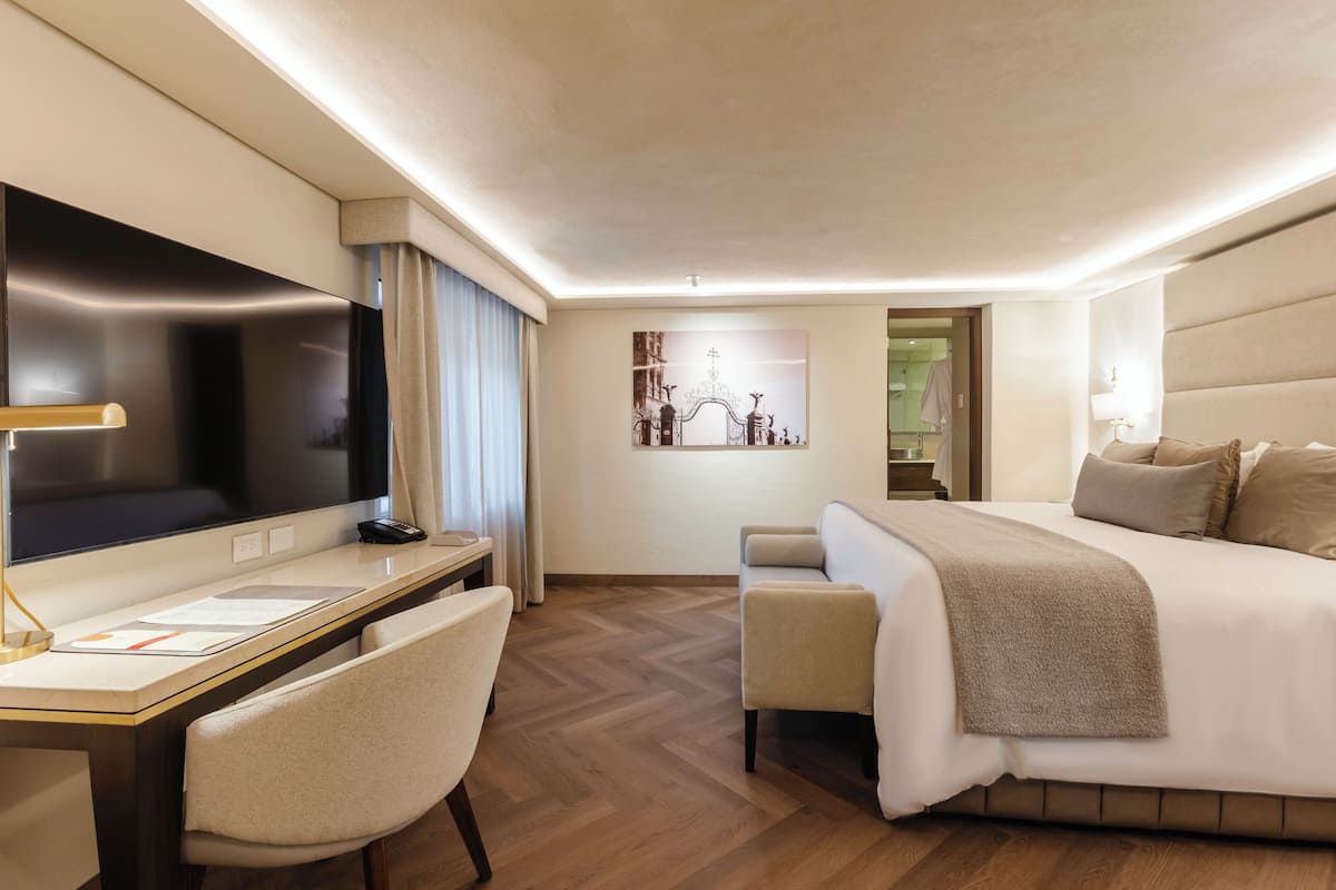Une chambre d’hôtel moderne avec un grand lit, un bureau doté d’un téléviseur à écran plat, une chaise et un tableau au mur.