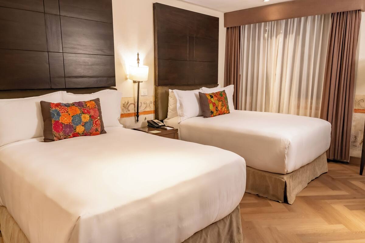 Une chambre d’hôtel avec deux lits, chacun agrémenté de draps blancs et d’oreillers aux motifs floraux colorés. On y trouve une table de chevet avec une lampe et un téléphone. La chambre est dotée d’un plancher en bois et d’une grande fenêtre avec des rideaux.