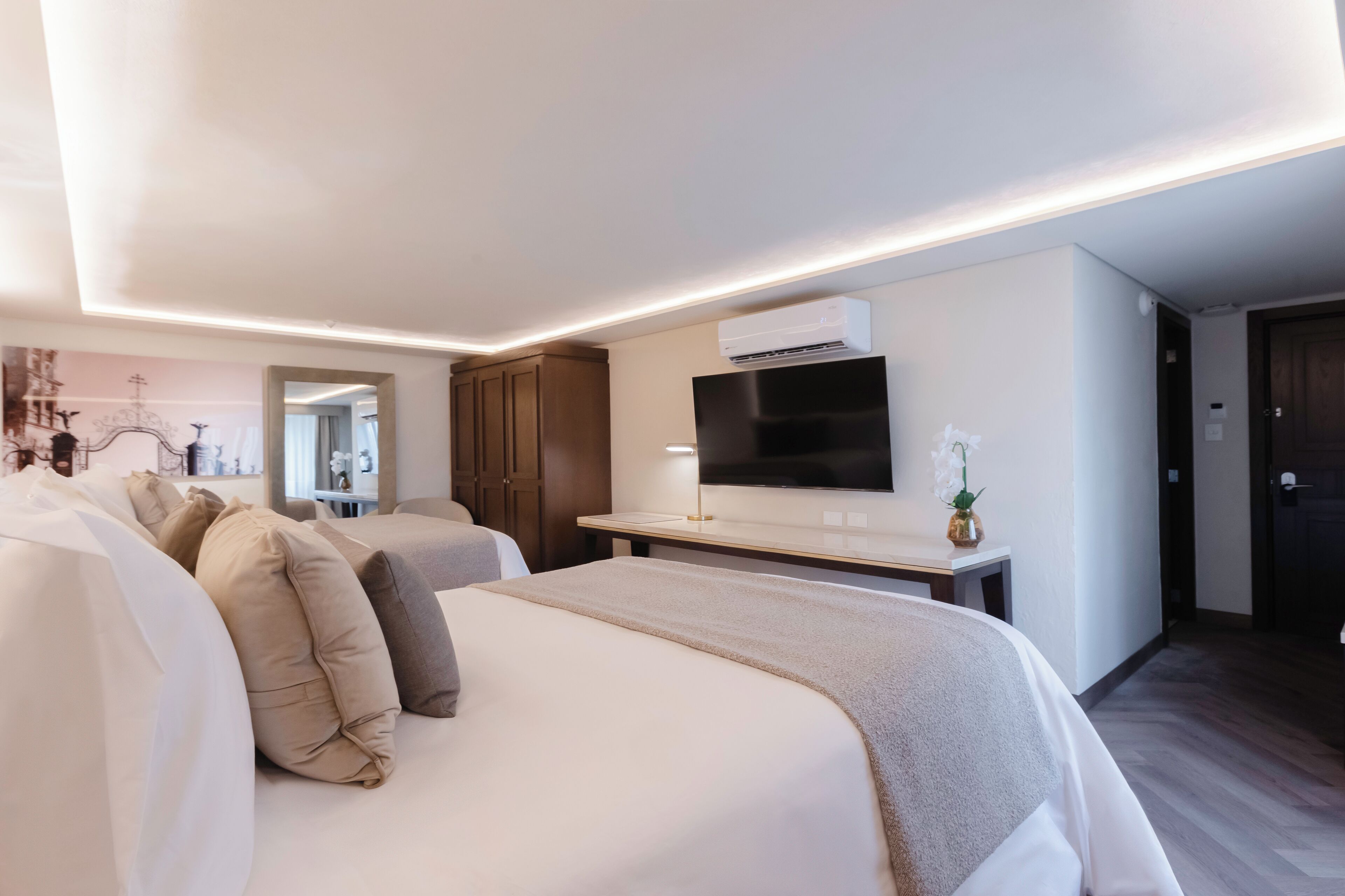 A modern hotel room with two beds, a flat-screen TV, an air conditioning unit, and a small table with a vase.