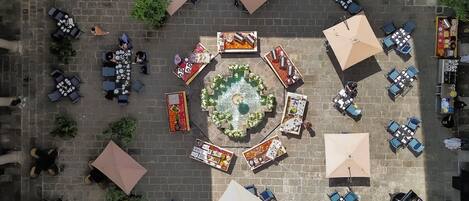 Vista aérea de un patio con mesas y sillas al aire libre, un jardín central y una fuente.