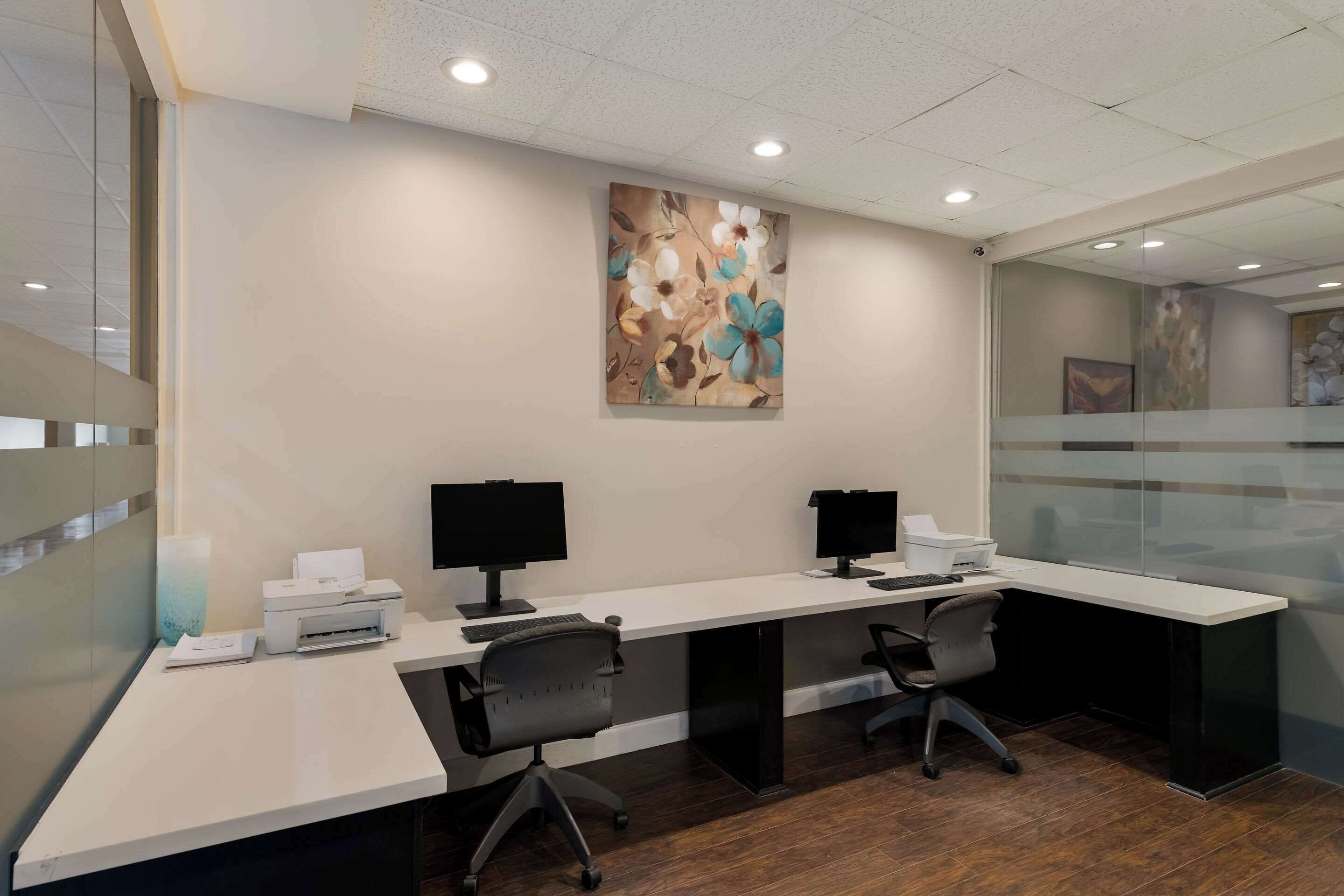 A modern office with two desks, each with a computer and chair, and a large floral painting on the wall.