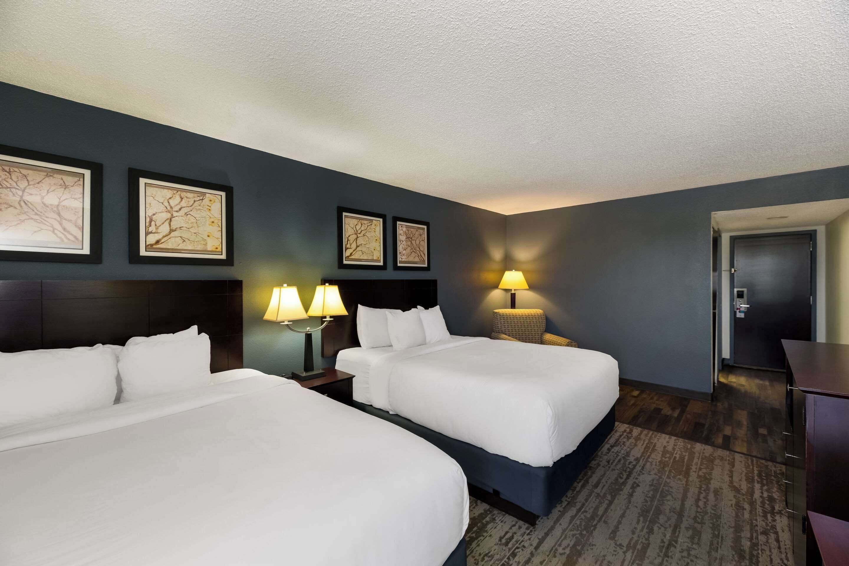 A hotel room with two beds, a desk, and a chair. There are framed pictures on the wall and a lamp on the desk.