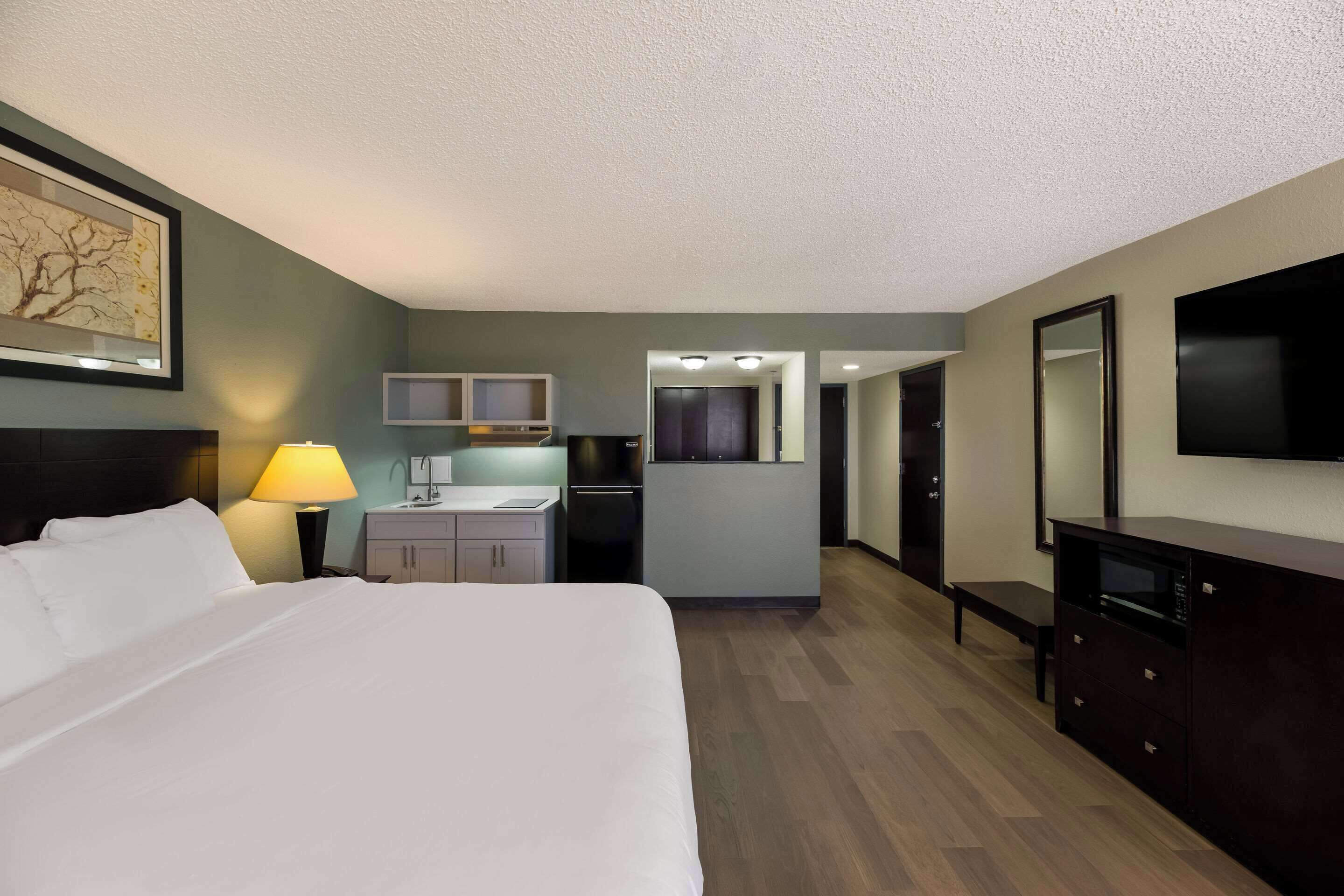 A hotel room with a bed, a desk, a kitchenette, and a TV.