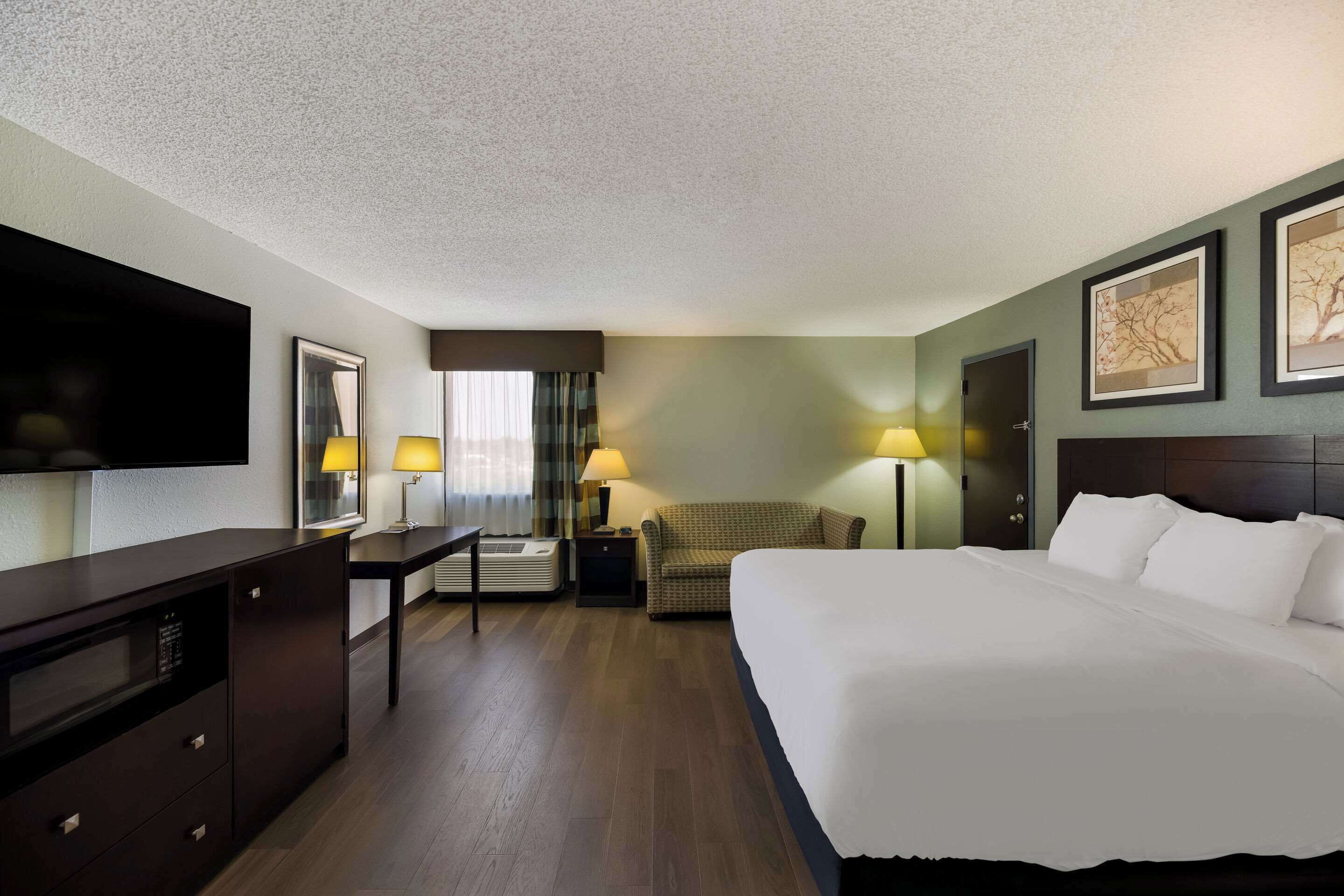 A hotel room with a large bed, a desk with a flat-screen TV, two bedside lamps, a sofa, and a door.