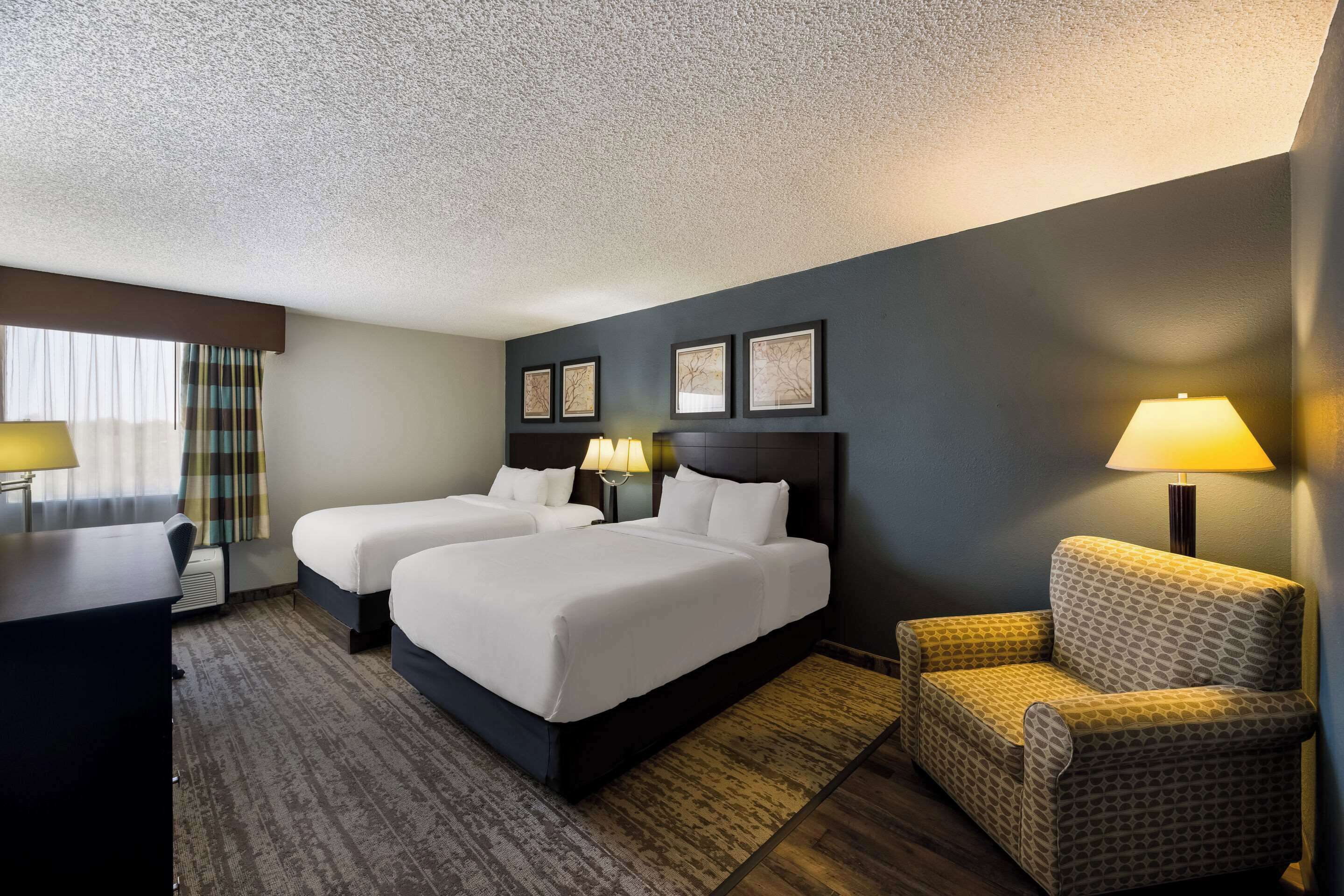A hotel room with two beds, a desk, a chair, and a lamp.
