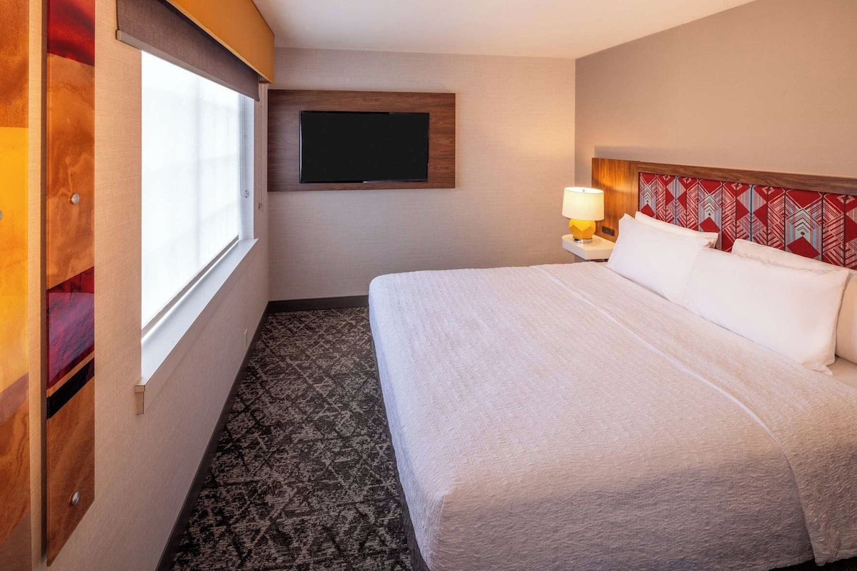 Hotellihuone, jossa on sänky, seinälle asennettu televisio, yöpöytä lampun kanssa ja kuviollinen matto.