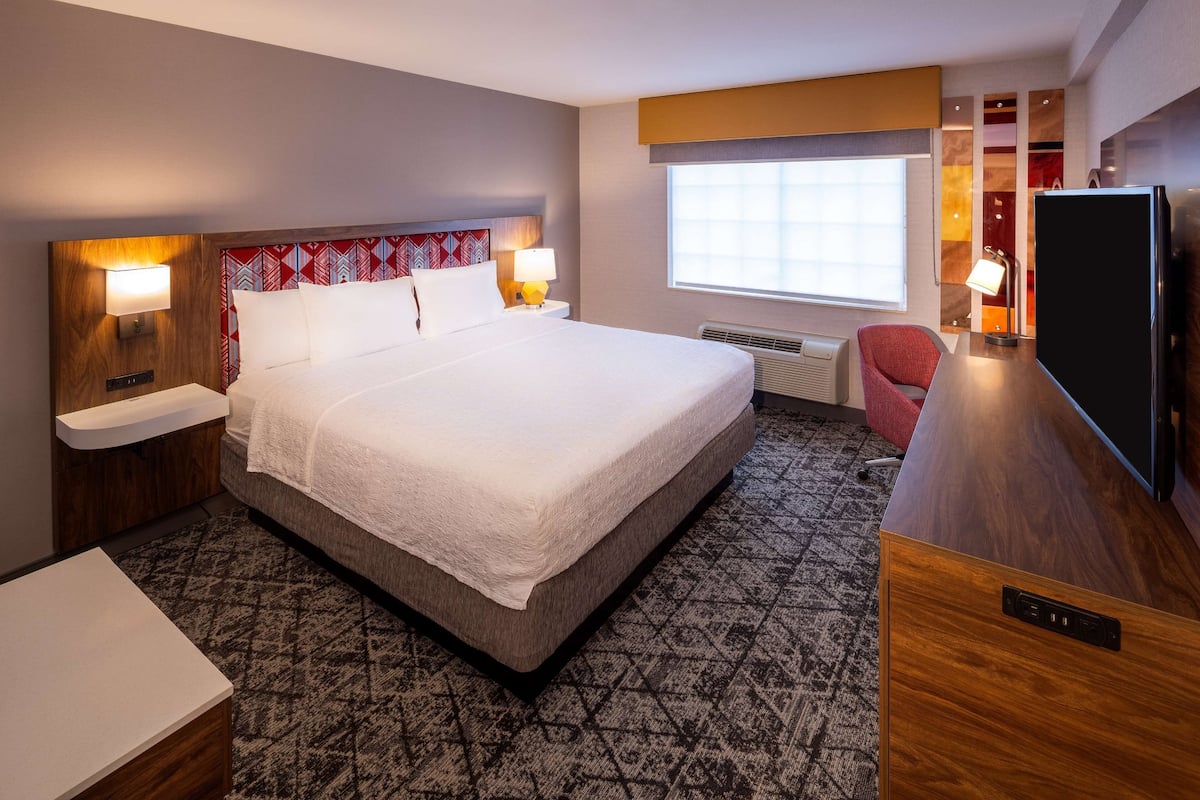 Quarto de hotel com cama grande, abajures, televisão, secretária e janela com persianas.