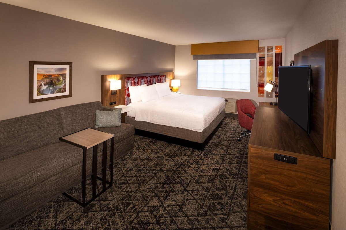 Quarto de hotel com cama, sofá, mesa pequena, televisão e janela.