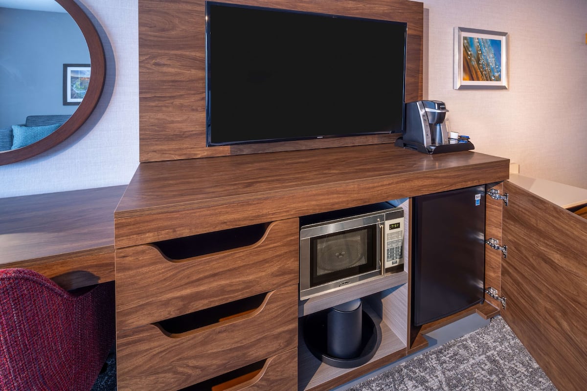 Quarto de hotel moderno com unidade de entretenimento em madeira, televisão de tela plana, cafeteira, micro-ondas e lixeira.