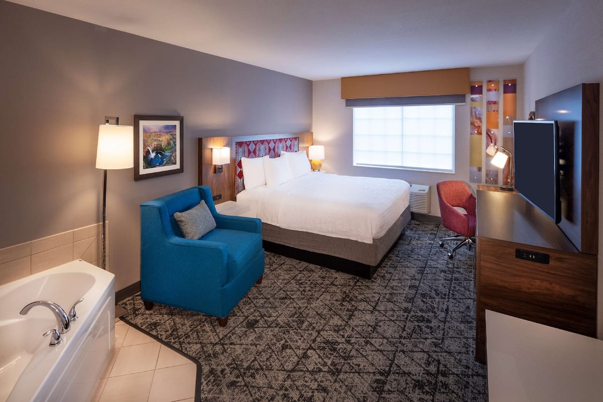 Quarto de hotel com uma cama grande, uma poltrona azul, uma banheira branca e uma televisão.