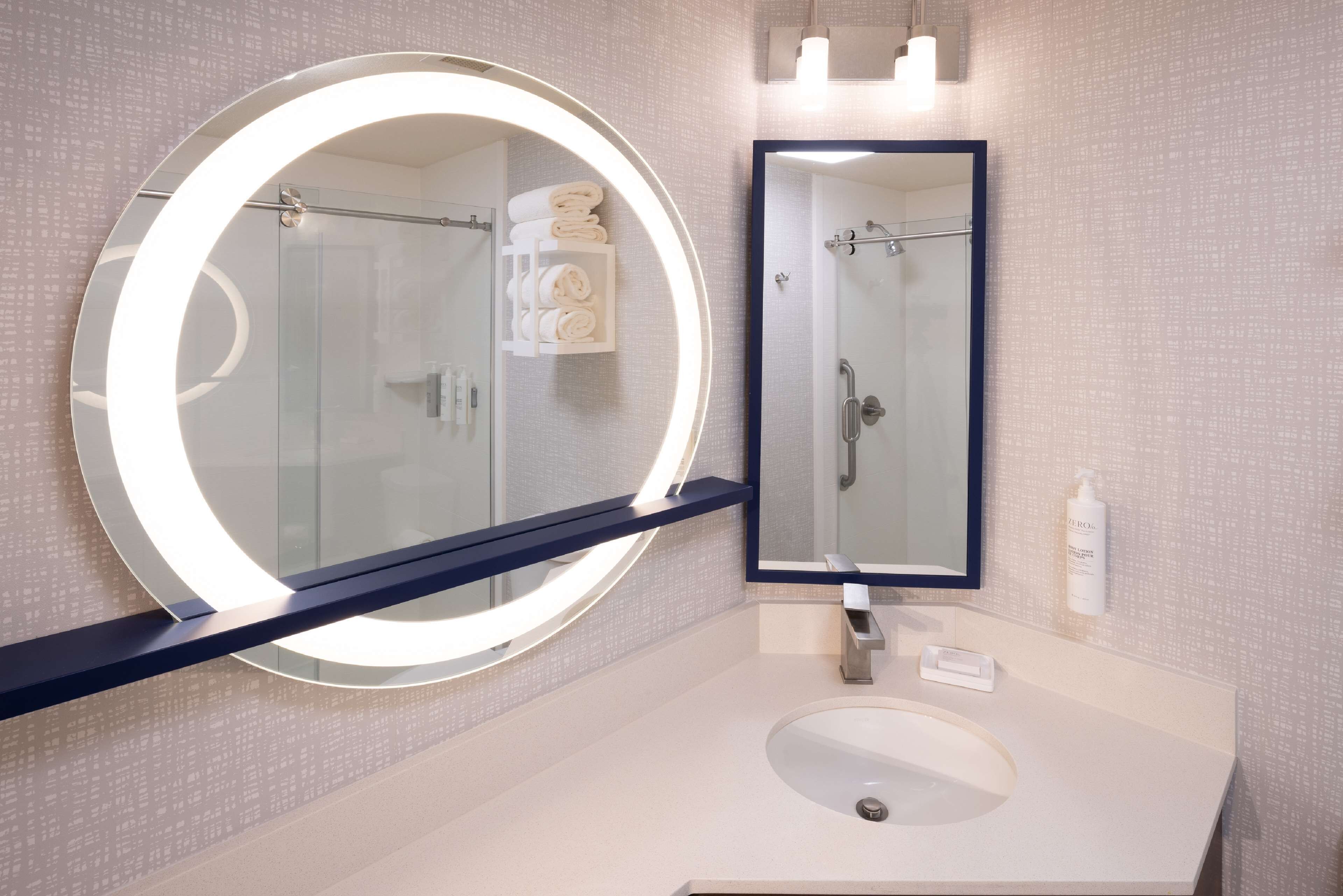 Een moderne badkamer met een ronde spiegel, een rechthoekige spiegel, een witte wasbak en een douchecabine met glazen wanden.