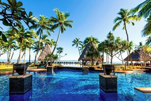 A pool area with palm trees, a thatched-roof structure, and a view of the ocean.